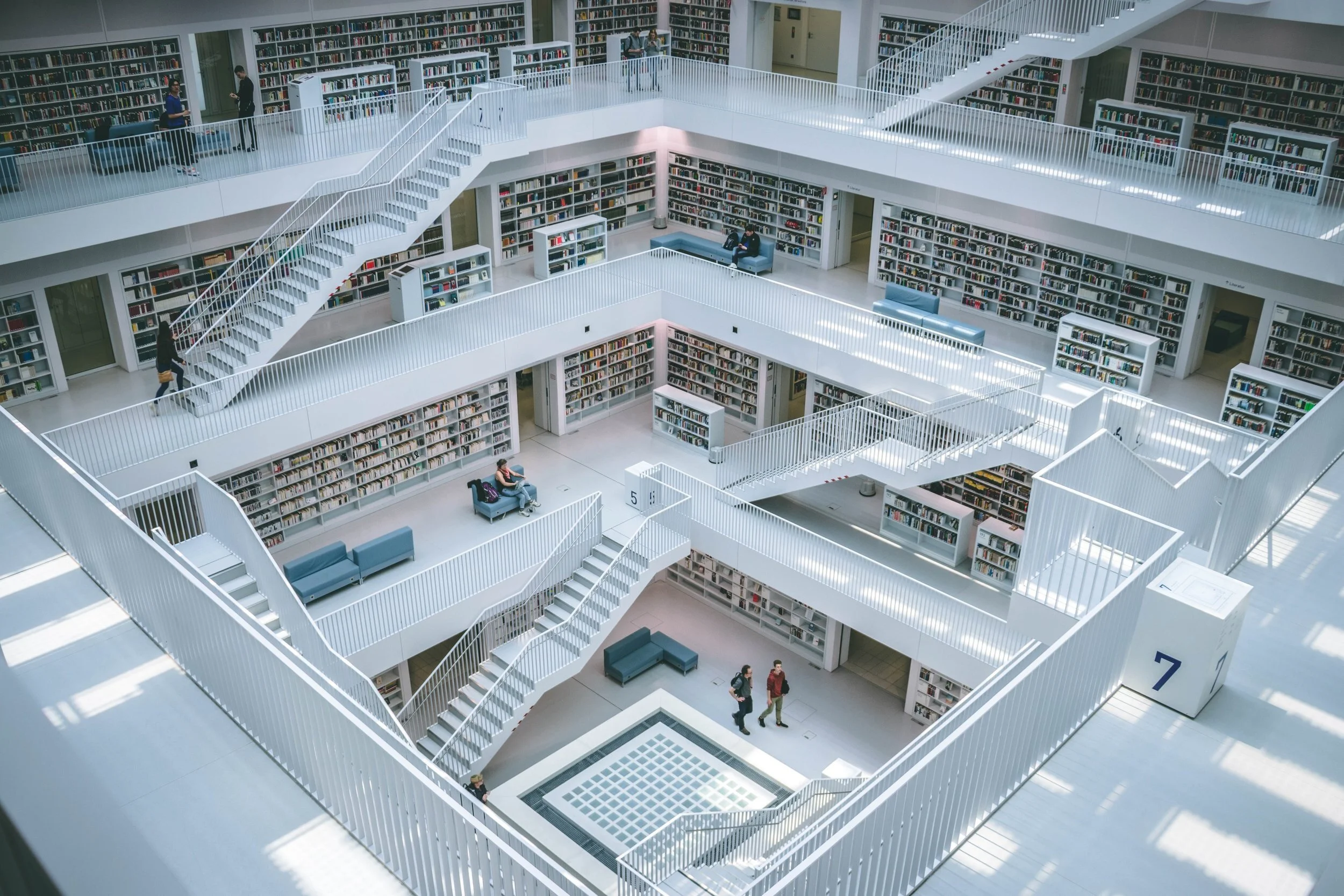 Multiple levels of a modern library with white railings and bookshelves filled with books. People are walking, reading, and sitting on benches. The architecture features a central open space with staircases connecting the floors.
