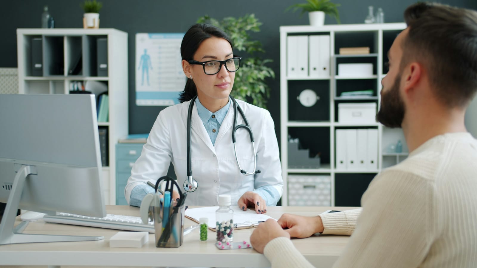 Clinician in a white coat consulting with a patient across a desk in a medical office