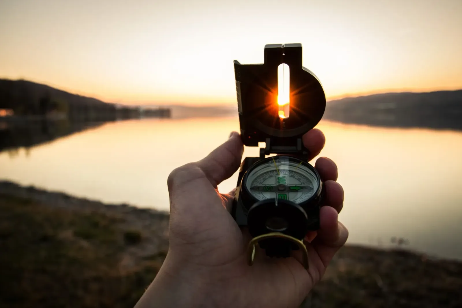 A hand holding a compass beside water at sunset.