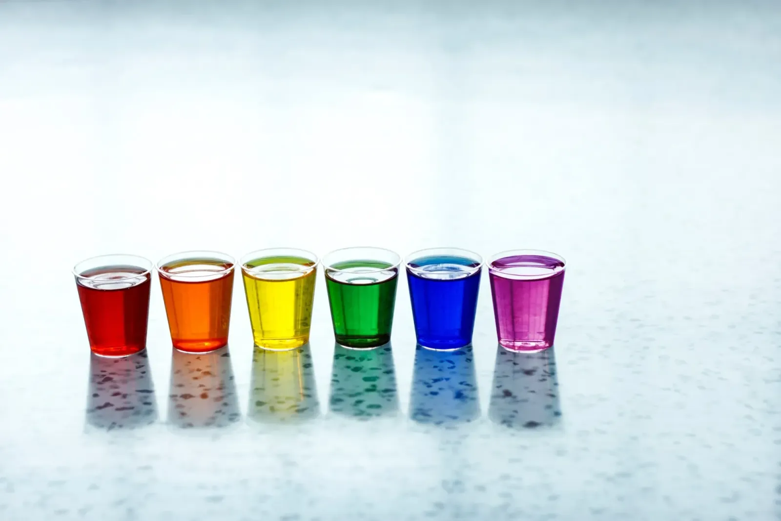 Six glasses filled with rainbow-colored liquids on a pale surface.