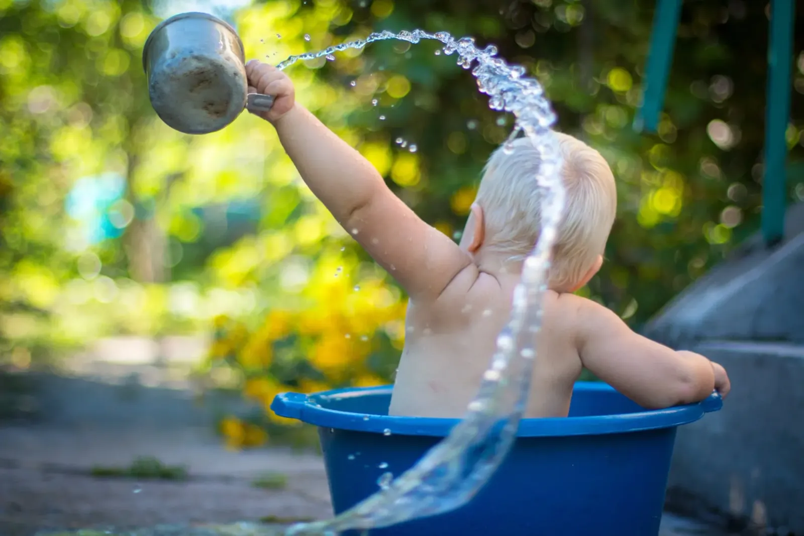 A young child splashing water from a metal cup while sitting in a blue tub outdoors.